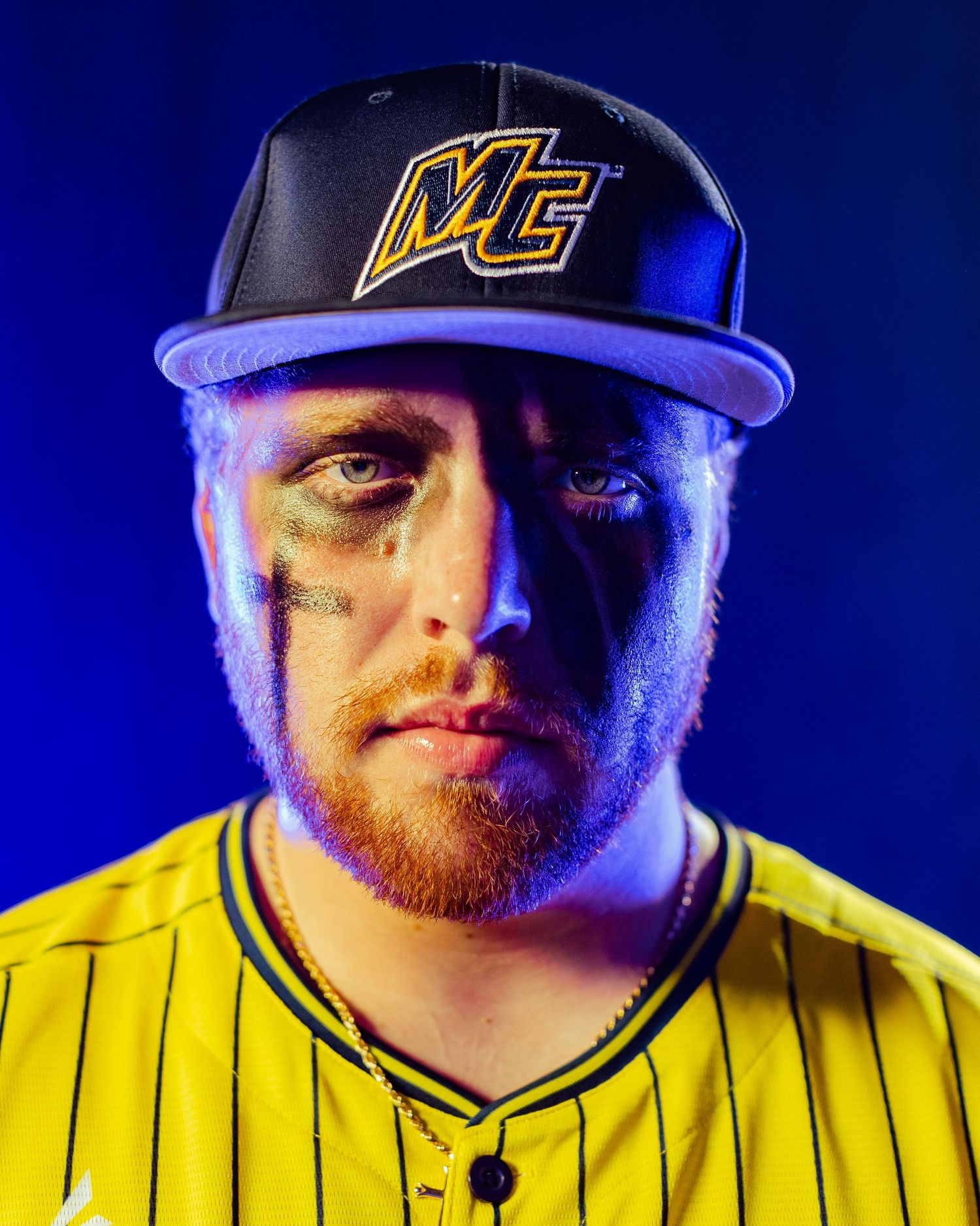 Baseball player in yellow pinstripe jersey and black MC cap with eye black, dramatic blue studio lighting.