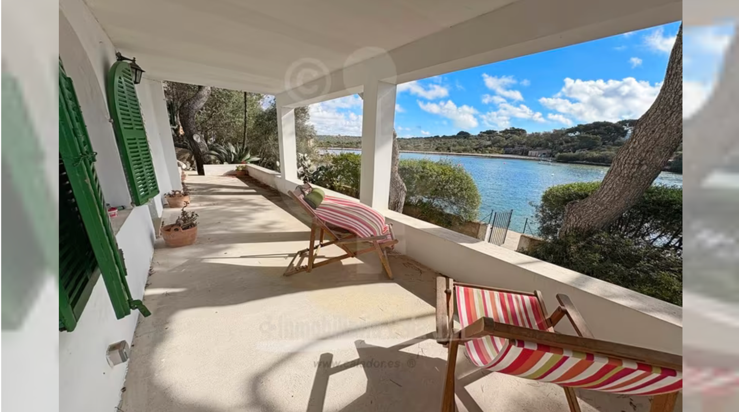 A waterfront porch with red and white striped lounge chairs overlooks a serene bay under blue skies.
