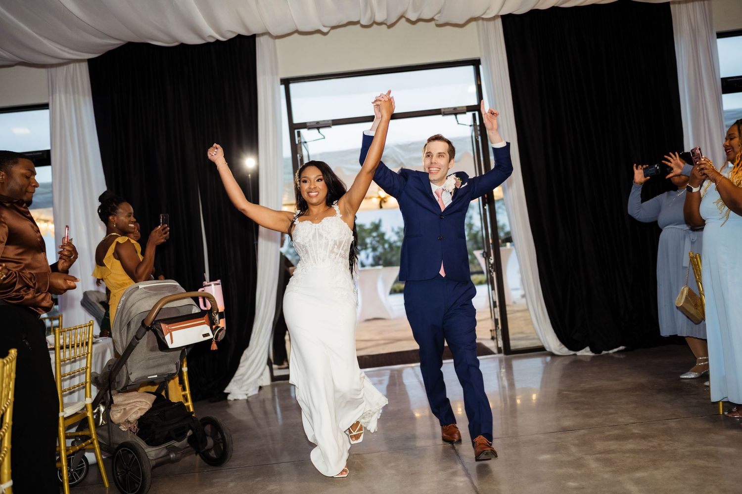 Couple celebrates their wedding entrance with raised arms while guests cheer during indoor reception.