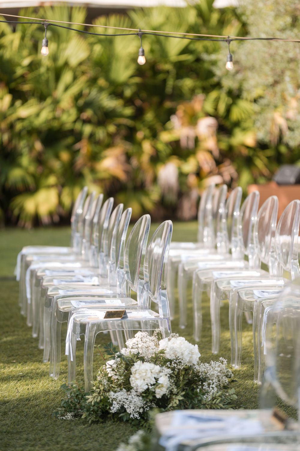 Outdoor wedding ceremony setup with white chairs aligned on the grass and floral arrangements along the aisle