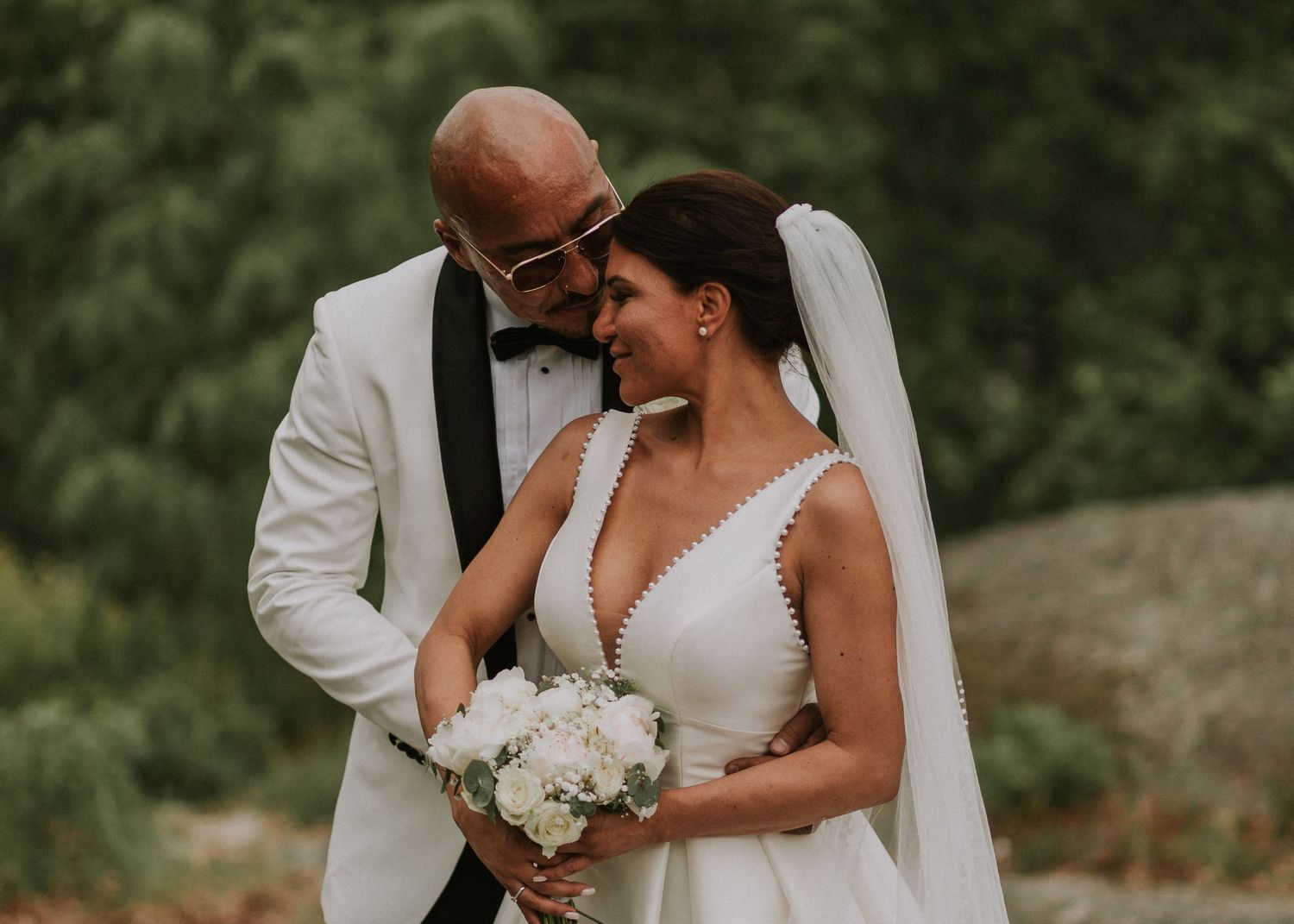Bride and groom share a tender moment, with groom kissing brides forehead outdoors.