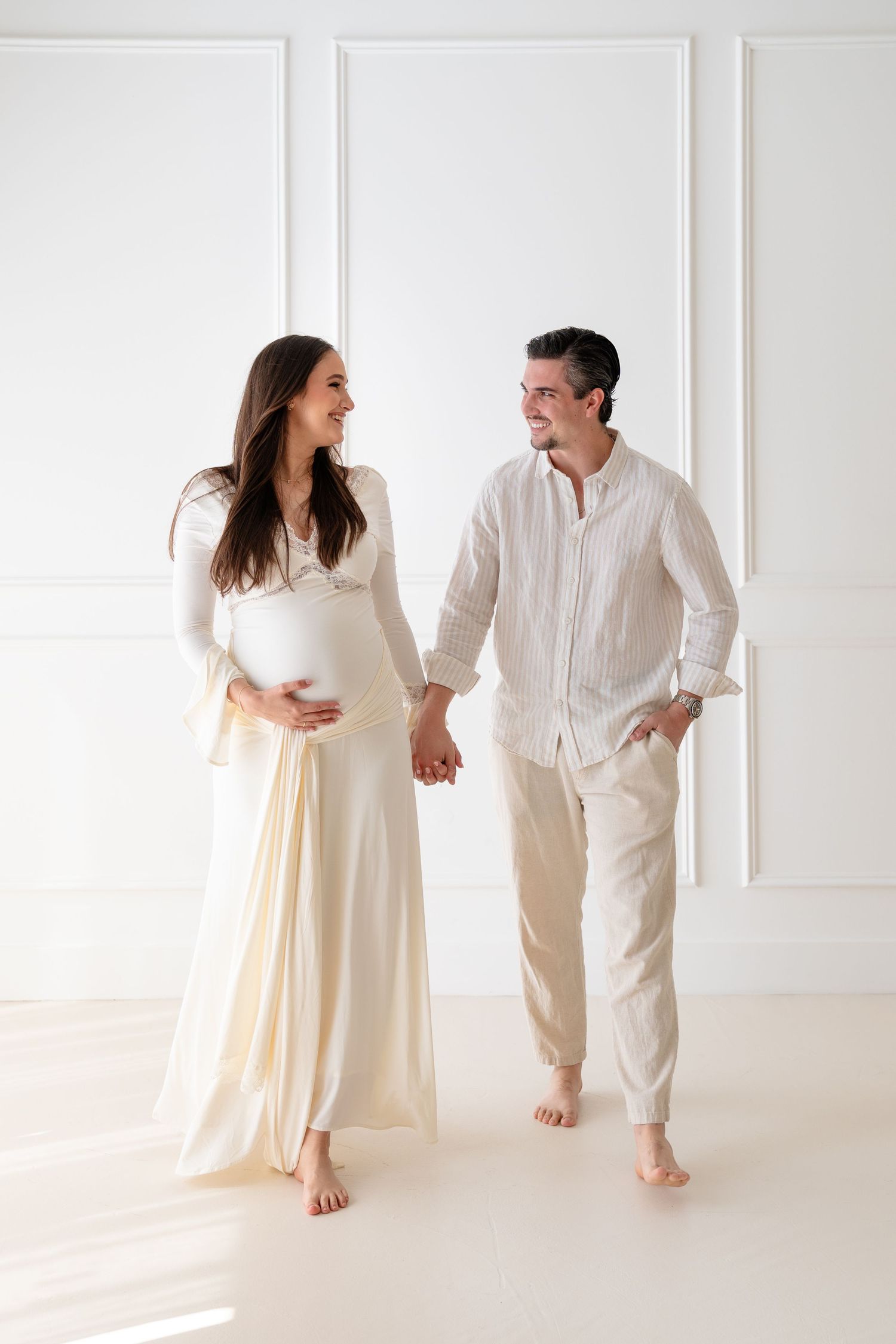 Couple in white outfits dancing together in bright minimalist studio with natural light streaming through windows.