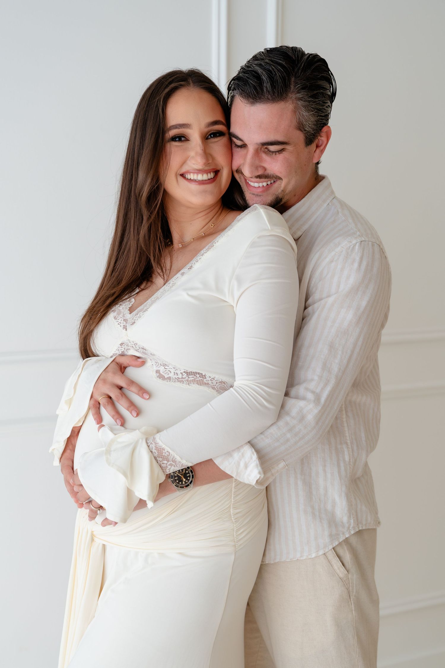 A couple in white outfits embraces during a maternity photoshoot against a light-colored wall.