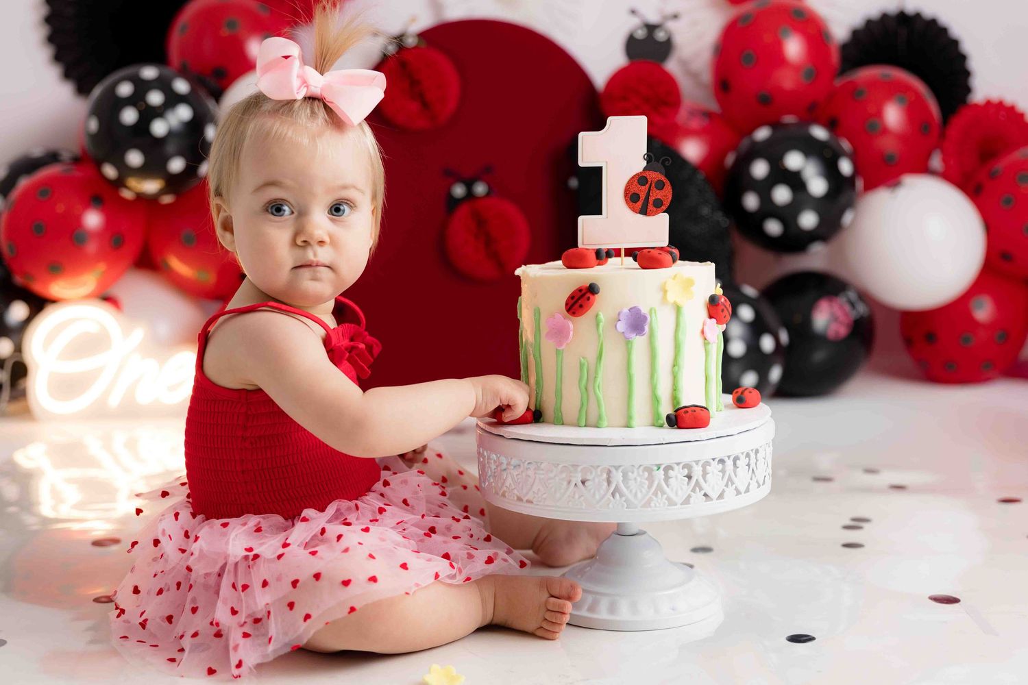 Baby in red tutu dress sits among red and black balloons during festive photo session.