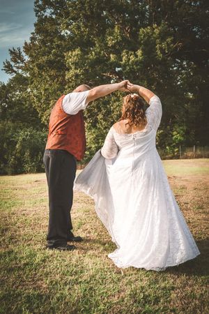 A couple in wedding attire dance together in a grassy field with trees in the background during golden hour.
