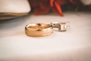 A gold wedding band with intricate details sits on a soft white surface with a blurred background.