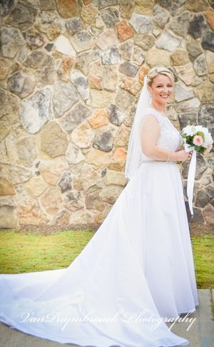 A flowing white wedding dress with a long train sweeps across the grass against a rustic stone wall backdrop.