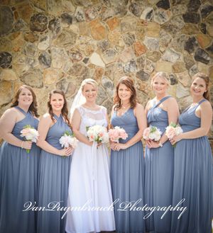 Wedding party in dusty blue bridesmaid dresses and white bridal gown pose in front of rustic stone wall.