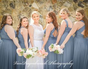 A row of bridesmaids in dusty blue dresses stand together holding pink and white bouquets at an outdoor wedding.