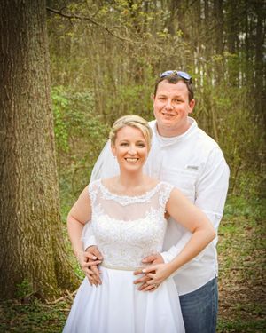 A couple embraces in a wooded outdoor setting with the person in a white dress smiling warmly.