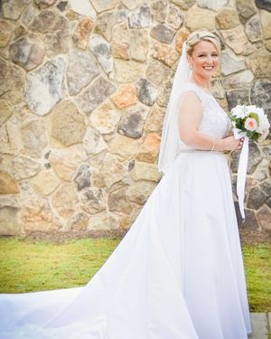 A bride in an elegant white gown with flowing train poses against a rustic stone wall outdoors.