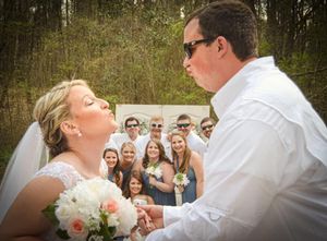 Wedding couple shares moment while guests react in background near wooded area.