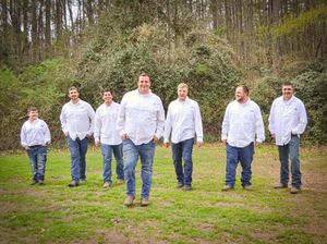 Group of men in white shirts and jeans pose on grassy hill for wedding party photo.