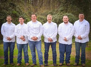 Six men wearing white shirts and blue jeans stand in a row outdoors against a wooded background.