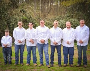 A row of groomsmen in white shirts and jeans pose casually outdoors against a natural background.