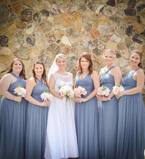 A bridal party in blue bridesmaid dresses and white wedding gown pose against a rustic stone wall.