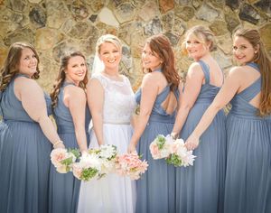 Bridesmaids in dusty blue dresses stand together holding pink and white floral bouquets at an outdoor wedding.