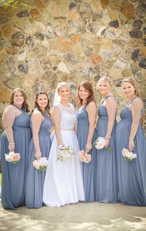 Bridesmaids in dusty blue dresses stand in line with bride in white, holding bouquets against stone wall backdrop.
