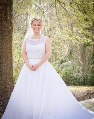 A person in a flowing white wedding dress poses outdoors in a wooded setting with natural sunlight filtering through trees.