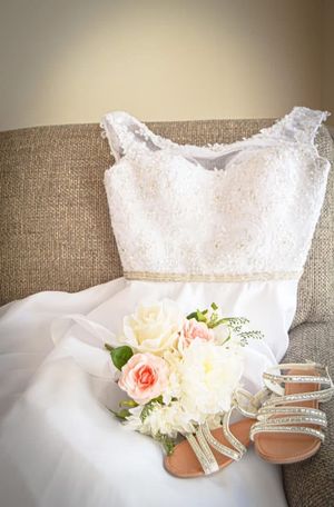Wedding dress, bouquet of pink and white roses, and sparkly sandals arranged on beige couch.