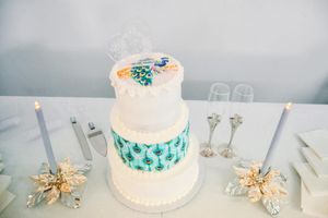 A white wedding cake with teal accents and gold decorative elements sits on display with glassware in the background.