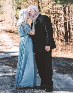 A couple in formal wear stands together on a snowy path surrounded by winter trees.