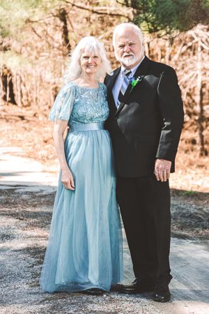 A couple in formal attire, with the woman wearing a light blue gown and the man in a black suit, pose outdoors.