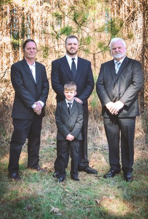 A group of formally dressed people stand in a line outdoors against a wooded backdrop in matching dark suits.