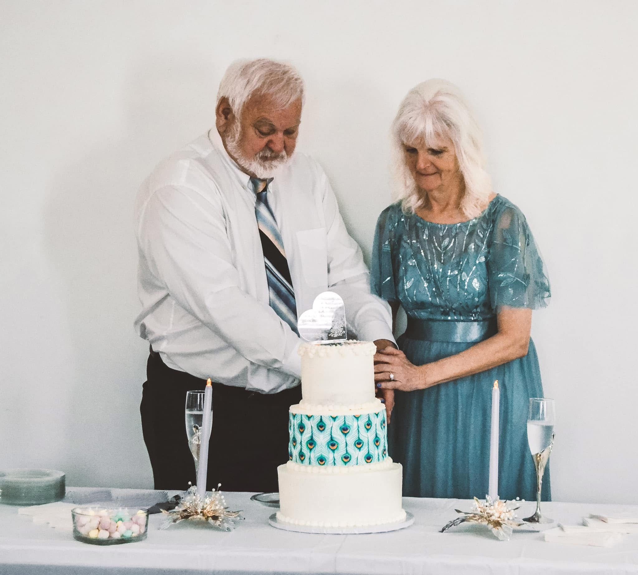 A couple in formal attire cuts into their elegant white wedding cake at a reception celebration.