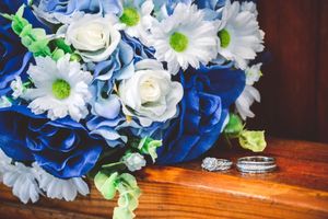 Wedding bouquet with blue roses and white daisies next to silver wedding rings on wooden surface.