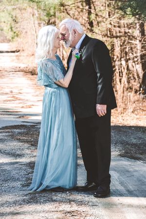 A couple in formal attire shares a romantic moment on a wooded path, with the person in a light blue dress.