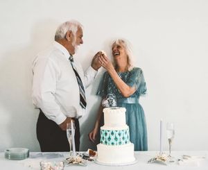 A couple in formal wear shares a playful moment while cutting their white and teal wedding cake together.