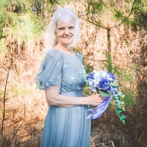 A person in a dusty blue dress holds a bouquet of blue and purple flowers against a sunlit wooded background.