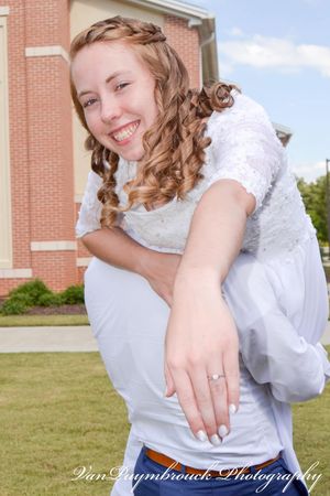A smiling figure in a white lace top and jeans sits casually outdoors near a brick building, showing off curled hair and jewelry.