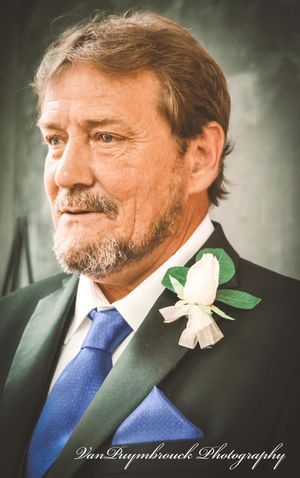 A formal portrait of a man in a dark suit with blue tie and white boutonniere at what appears to be a wedding event.