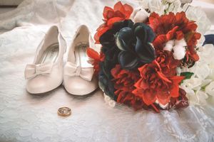 White bridal shoes and wedding rings displayed next to a dramatic red and black floral bouquet on white fabric.