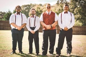 A group of groomsmen in suspenders and bowties stand together outdoors during a rustic wedding celebration.