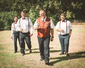A group in suspenders and vests walk together on a dirt path during an outdoor event.