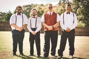 Groomsmen in suspenders and bow ties stand together in a rustic outdoor setting during a wedding celebration.