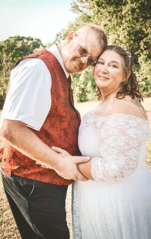 A couple in wedding attire poses outdoors, with the bride in a lace dress and groom wearing a red brocade vest.