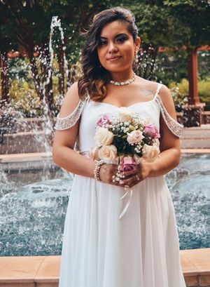 A bride in a flowing white dress holds a delicate bouquet while standing by a fountain in an outdoor garden setting.