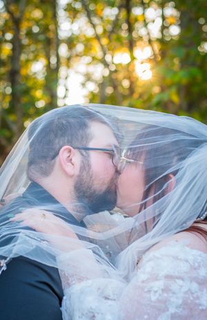 A couple shares an intimate kiss under a flowing white veil with sunlit autumn trees in the background.