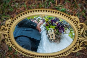 A wedding couple's reflection appears in an ornate gold mirror frame placed on fallen leaves outdoors.