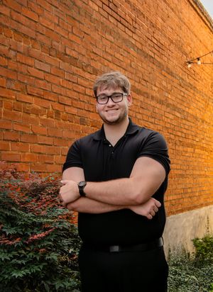 Person in a black polo shirt stands with crossed arms against an orange brick wall with green foliage at the base.