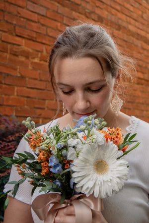 A series of four photos showing someone smelling a beautiful bouquet with white gerbera daisies and orange flowers against a brick wall.