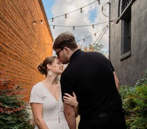 A couple shares an intimate moment against a brick wall with string lights and greenery in an outdoor setting.