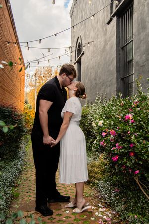 A couple shares a romantic moment in an alley with string lights and pink roses against brick and stone walls.