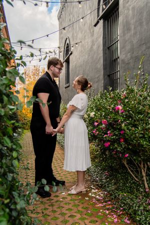 A couple shares an intimate moment in a garden setting with string lights and pink flowers against a concrete wall.