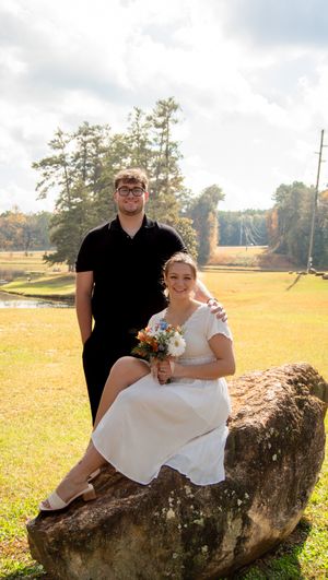 Couple poses together on large rustic log in sunny autumn field during outdoor wedding photoshoot.