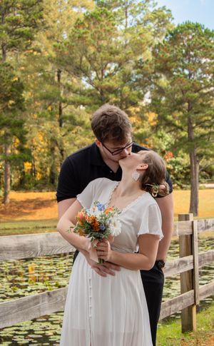 A romantic wedding moment captured by a pond with lily pads and autumn trees in the background.